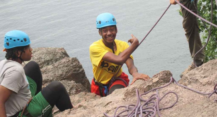 a person who is rock climbing smiles and points over their shoulder at the body of water below